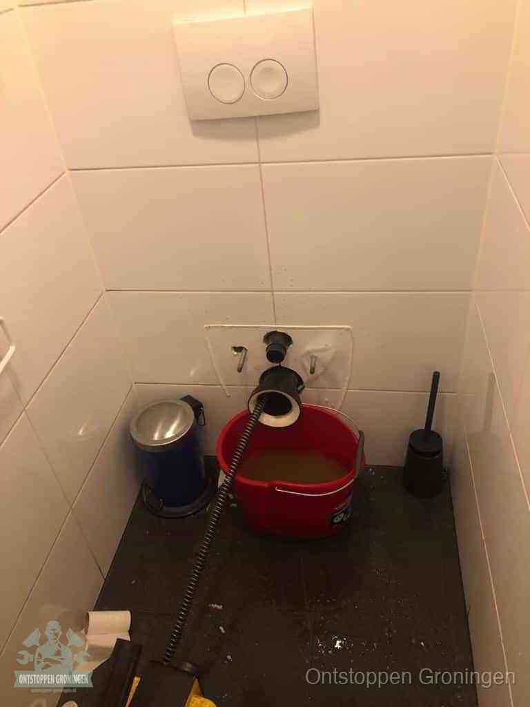 Een ontstopte wand-wc met verwijderde toiletpot waar een ontstoppingsveer in de wanduitgang verdwijnt, het achterhalen waar de verstopping zit in Groningen