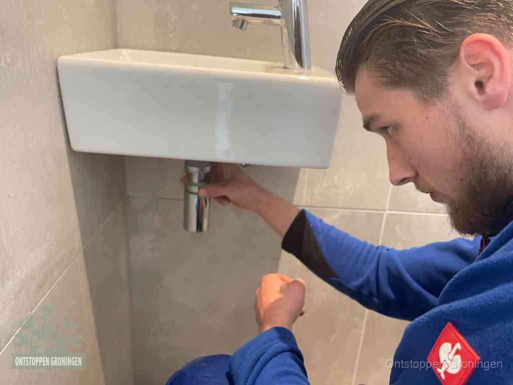 Een monteur draait met de hand de zwanenhalssifon onder een fonteinwasbak in de toiletruimte los om de bron van rioollucht in Groningen te vinden