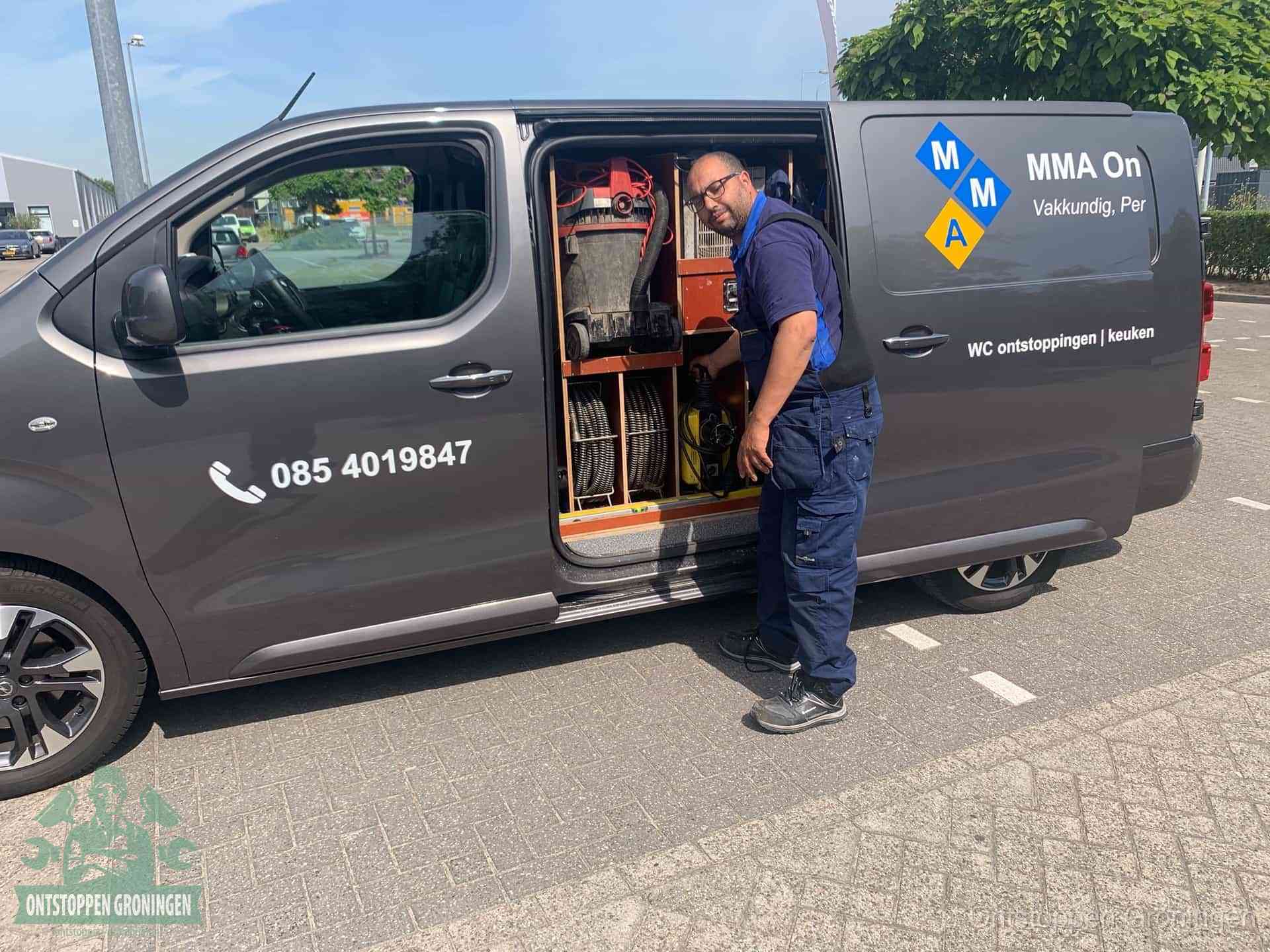 Een monteur bij de open schuifdeur van zijn bedrijfsbus op een parkeerplaats, klaar om de oorzaak van terugkerende afvoerverstoppingen aan te pakken in Groningen