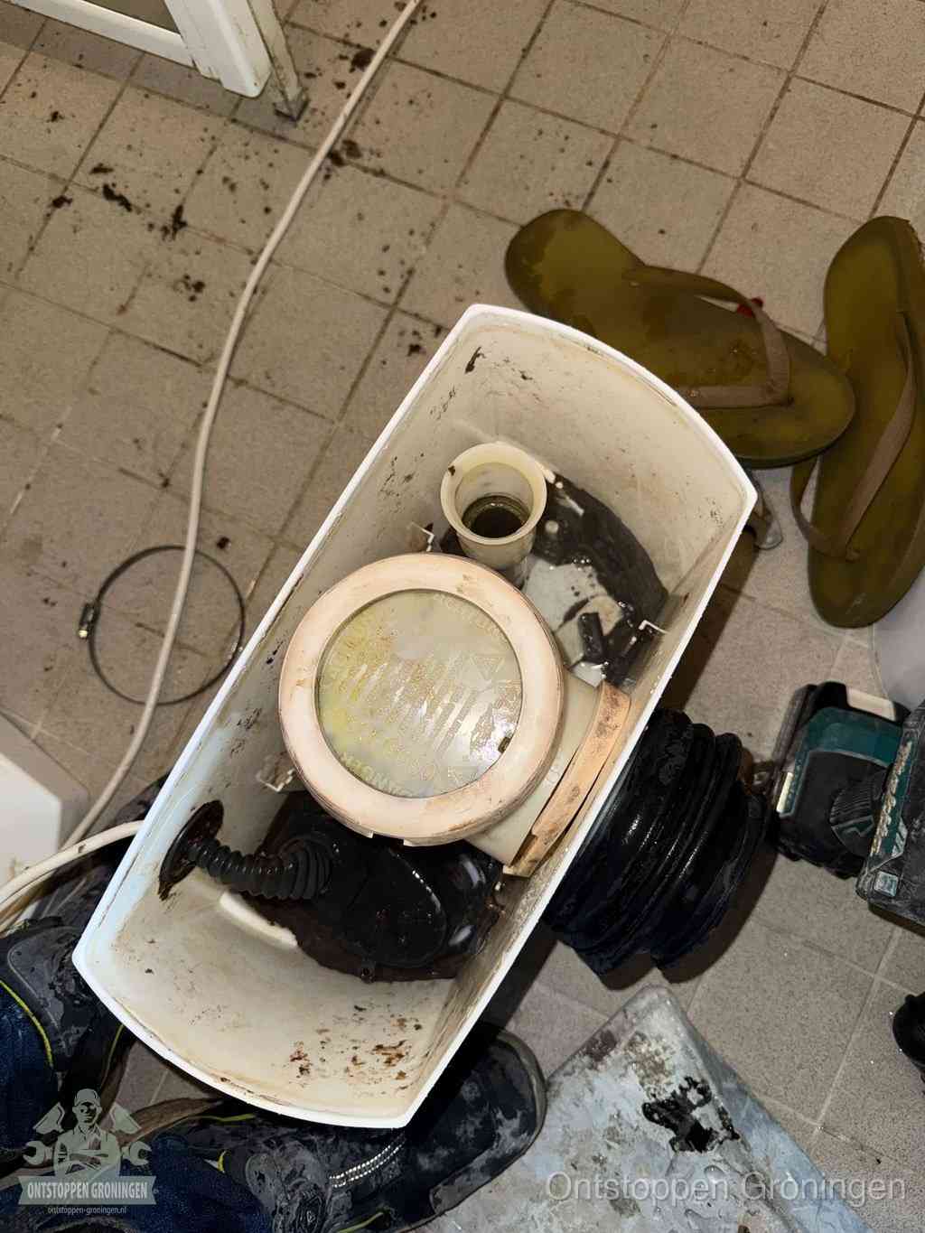 Een gedemonteerde toiletpot op de vloer met ontstoppingsveer en gereedschap ernaast, het verwijderen van een vastzittend voorwerp uit de wc in Groningen