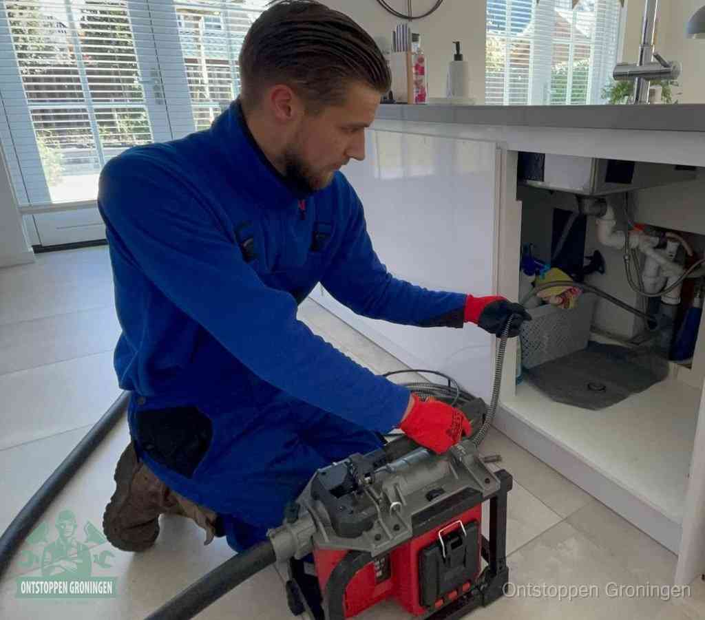 Een monteur met rode handschoenen knielt in een keuken en voert een elektrische ontstoppingsveer in tijdens het professioneel afvoer ontstoppen in Groningen